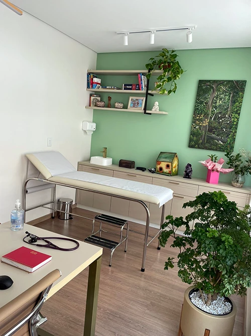 Sala de atendimento com maca de exame, estante de livros e plantas